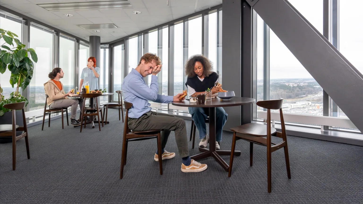 5. Fyra personer i ett modernt kontorsutrymme med stora fönster; två vid ett bord i förgrunden och två i bakgrunden vid en kaffestation.