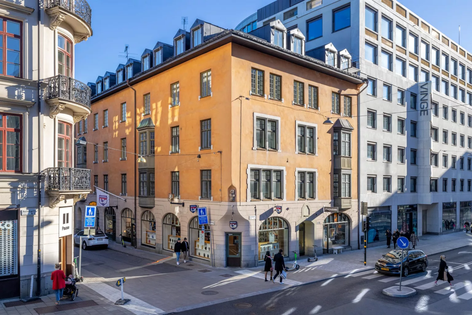 En gul byggnad med flera våningar, stora fönster och valv vid bottenvåningen, omgiven av andra moderna byggnader.