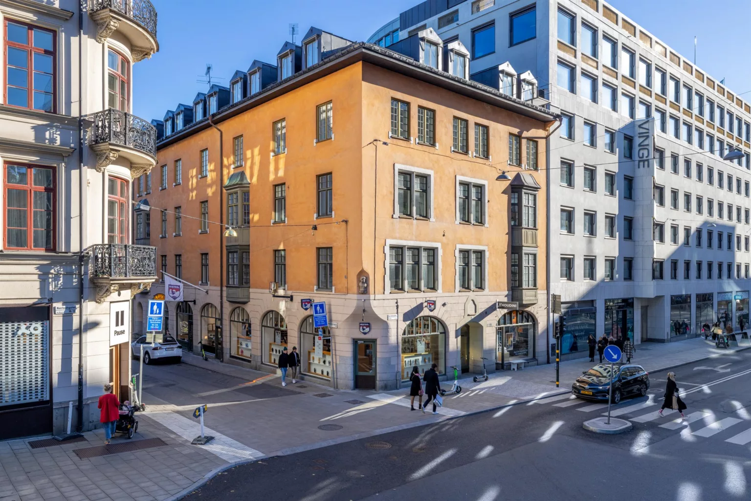 En gul byggnad med flera våningar och valv vid bottenvåningen, belägen vid en korsning med fotgängare och bilar.
