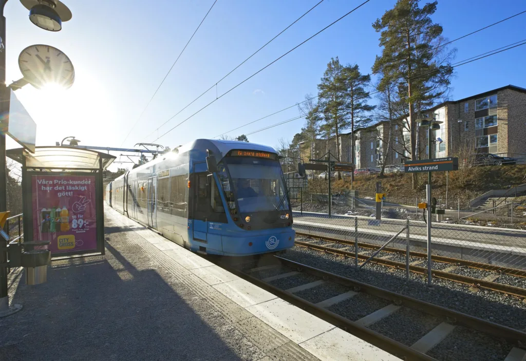 1. Ett blått pendeltåg står vid en perrong under en klarblå himmel med solen som lyser i bakgrunden.