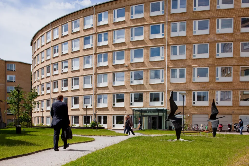 1. En rundad tegelbyggnad med många fönster och en grön gräsmatta framför, där människor går på en gångväg.