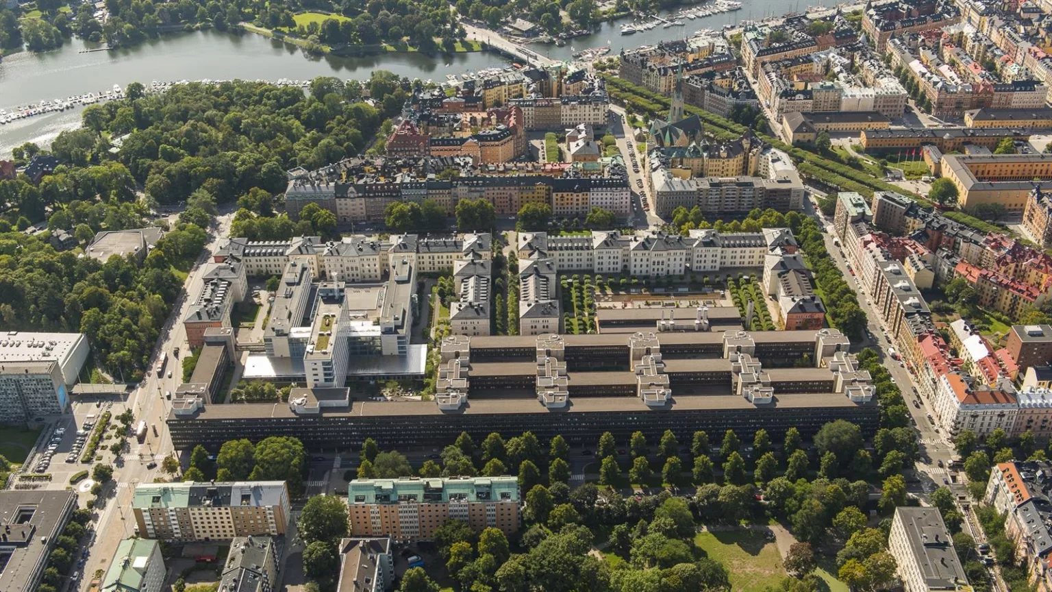 Flygbild av en stadsdel med regelbundna byggnader omgivna av grönområden och en flod i bakgrunden.
