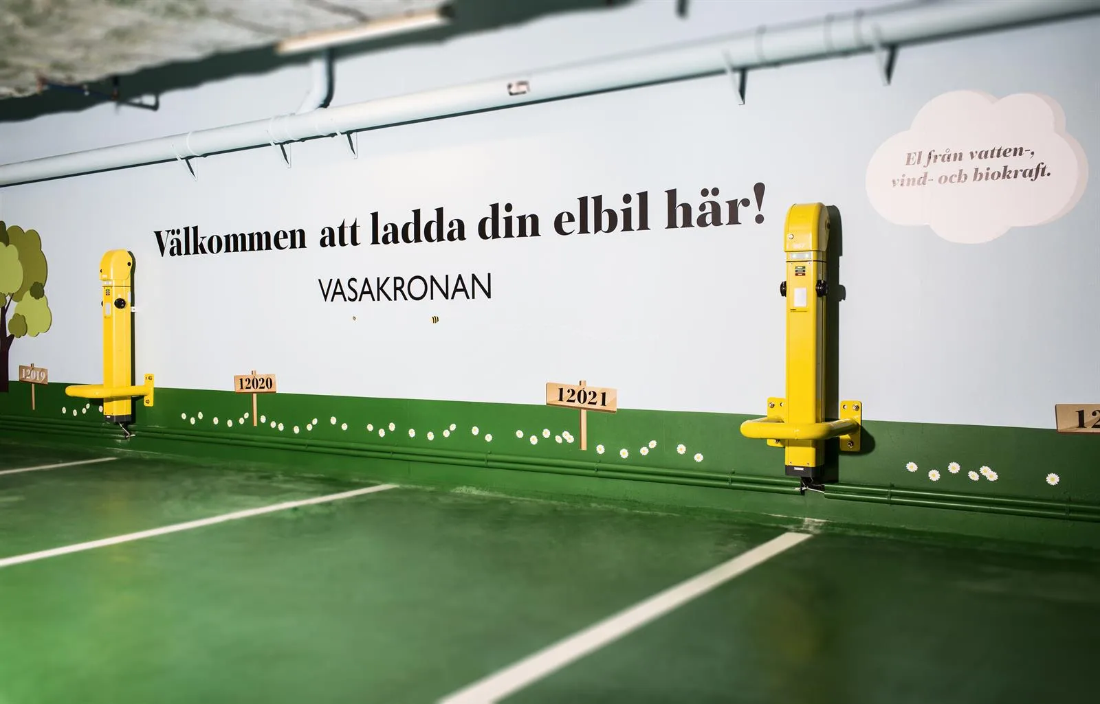 En gul laddningsstation för elbilar i ett grönt parkeringsområde med texten "Välkommen att ladda din elbil här! Vasakronan" på väggen.