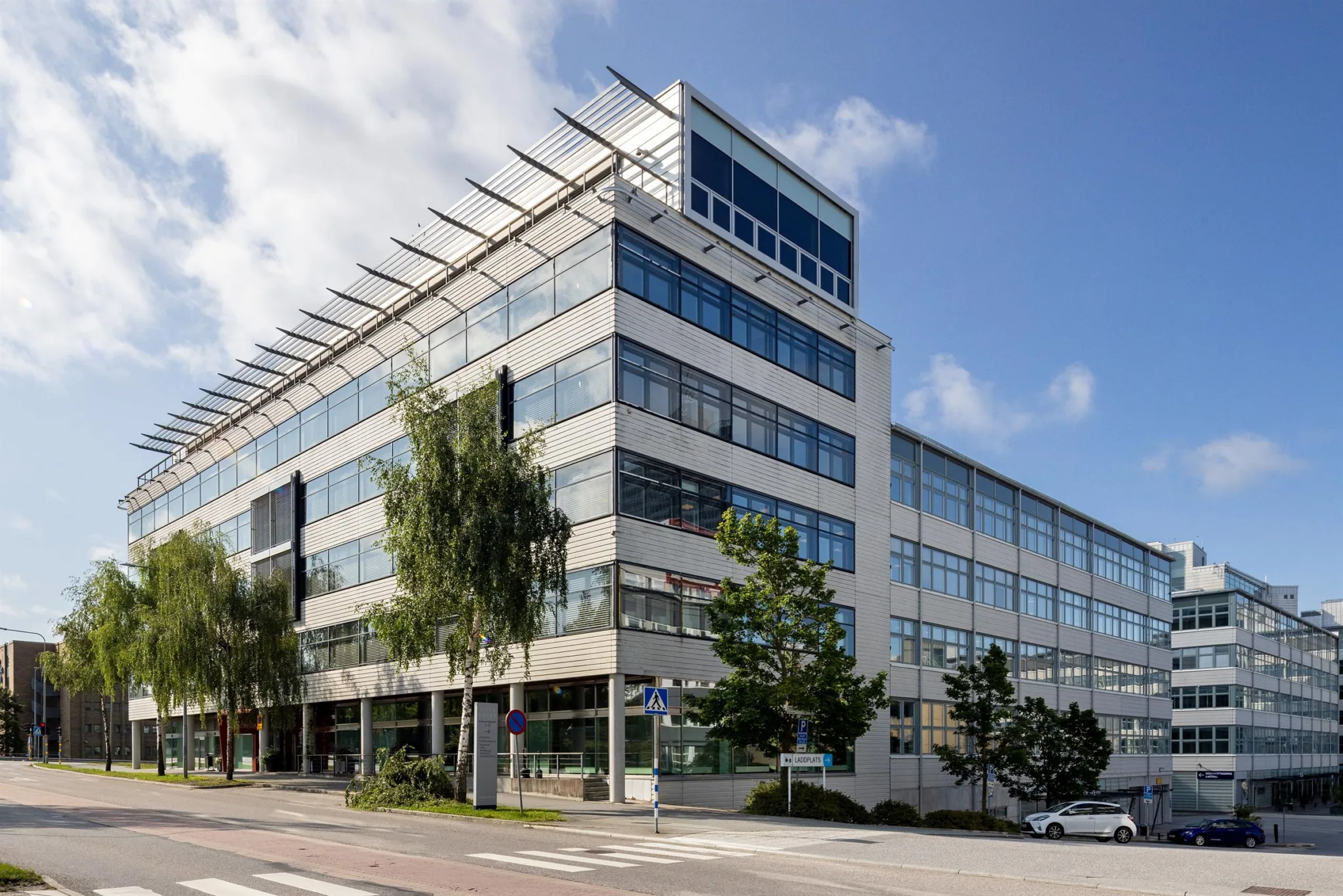 En stor modern kontorsbyggnad med glasfasad och flera våningar, omgiven av träd och en trottoar.