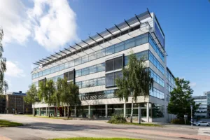 En modern byggnad med glasfasad och flera våningar, omgiven av träd och en trottoar.