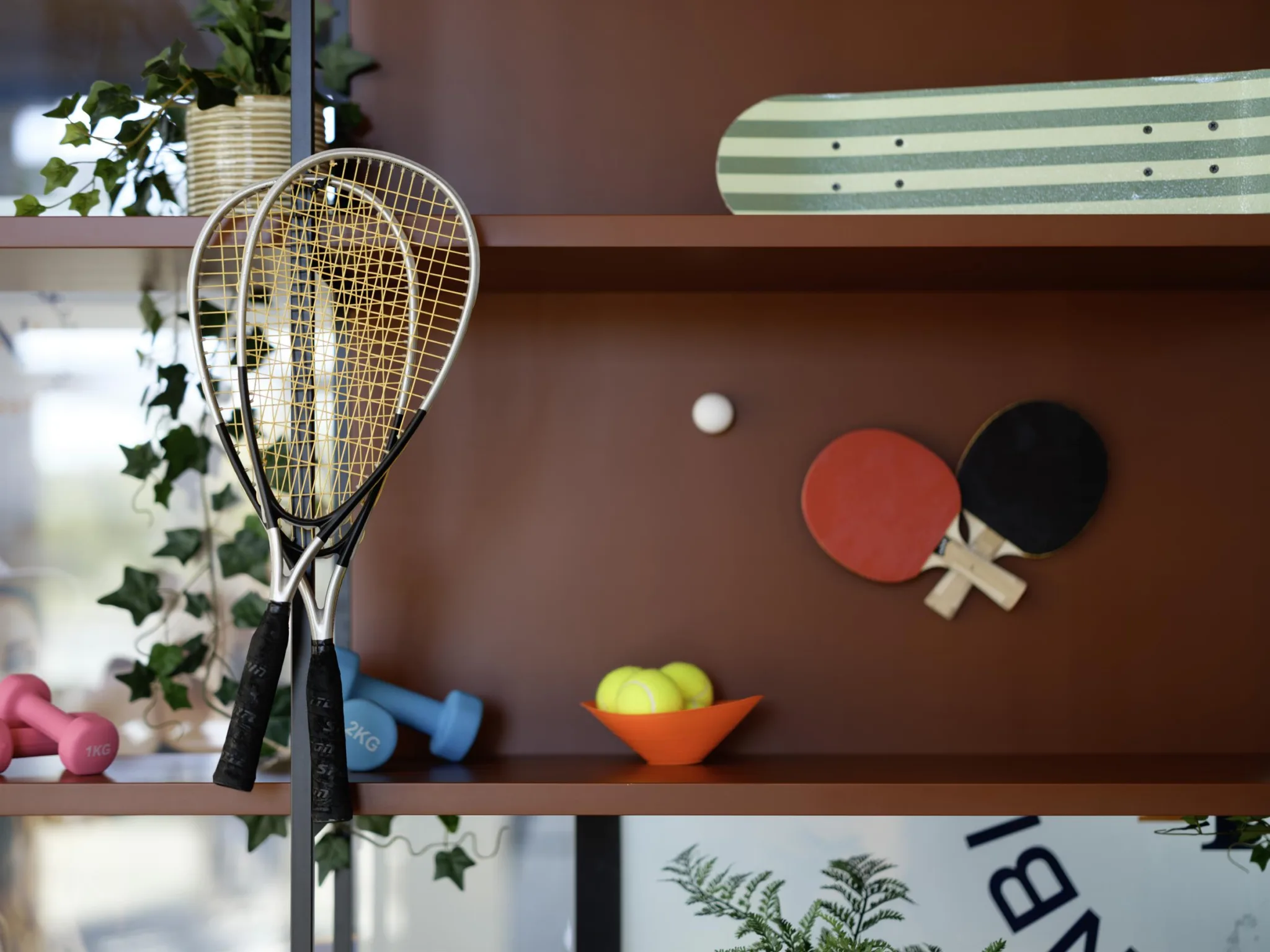 1. En hylla med sportutrustning, inklusive tennisracketar, pingisracketar och bollar, samt en skateboard och växter som dekoration.