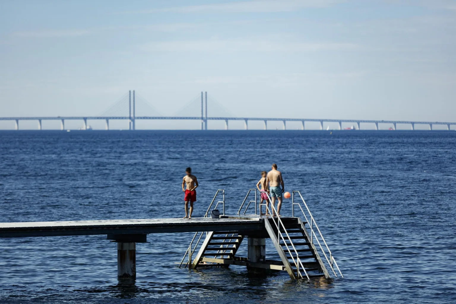 3. En brygga vid havet med människor som badar, och en bro syns i bakgrunden.