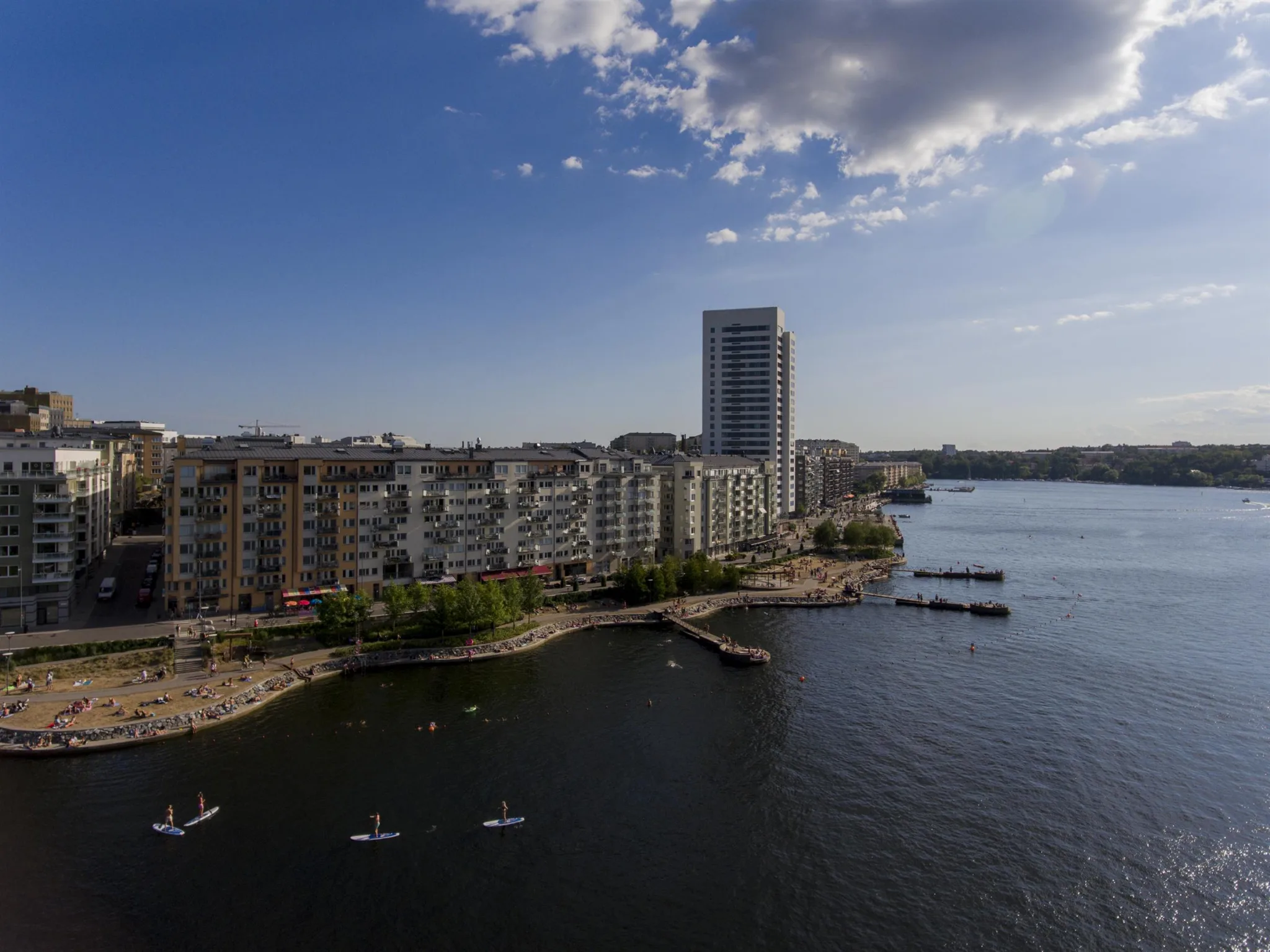 En stadsbild vid vattnet med höghus, bostäder och små båtar på det lugna vattnet under en blå himmel.