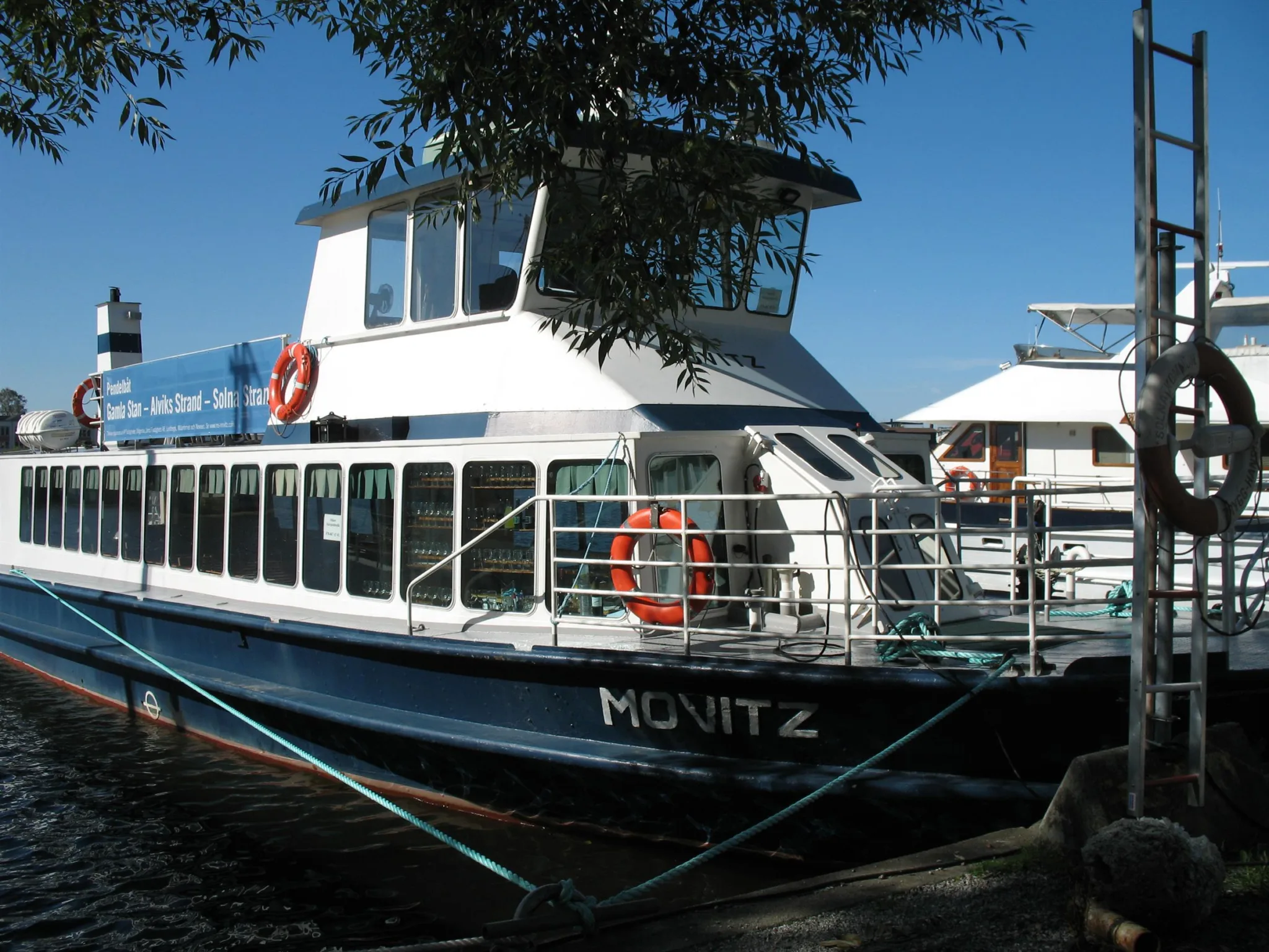 En båt med namnet "M/S Witz" ligger förtöjd vid en brygga, omgiven av träd och vatten.
