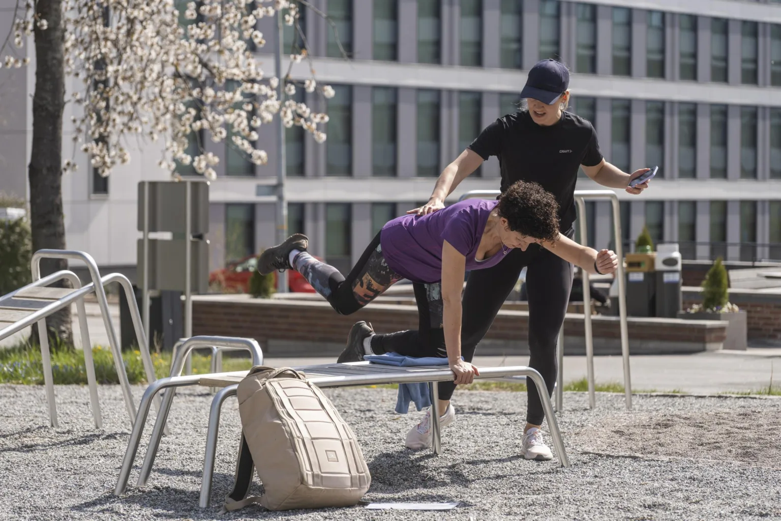 Två personer tränar utomhus på en hinderbana; en person hjälper den andra över en metallstång.