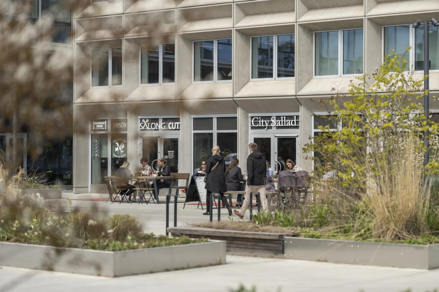 Människor som sitter och står utanför ett kafé vid en modern byggnad med stora fönster.