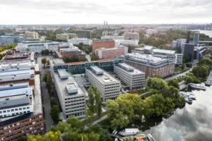 Flygvy över en stadsmiljö med moderna byggnader, grönska och en vattendrag i förgrunden.