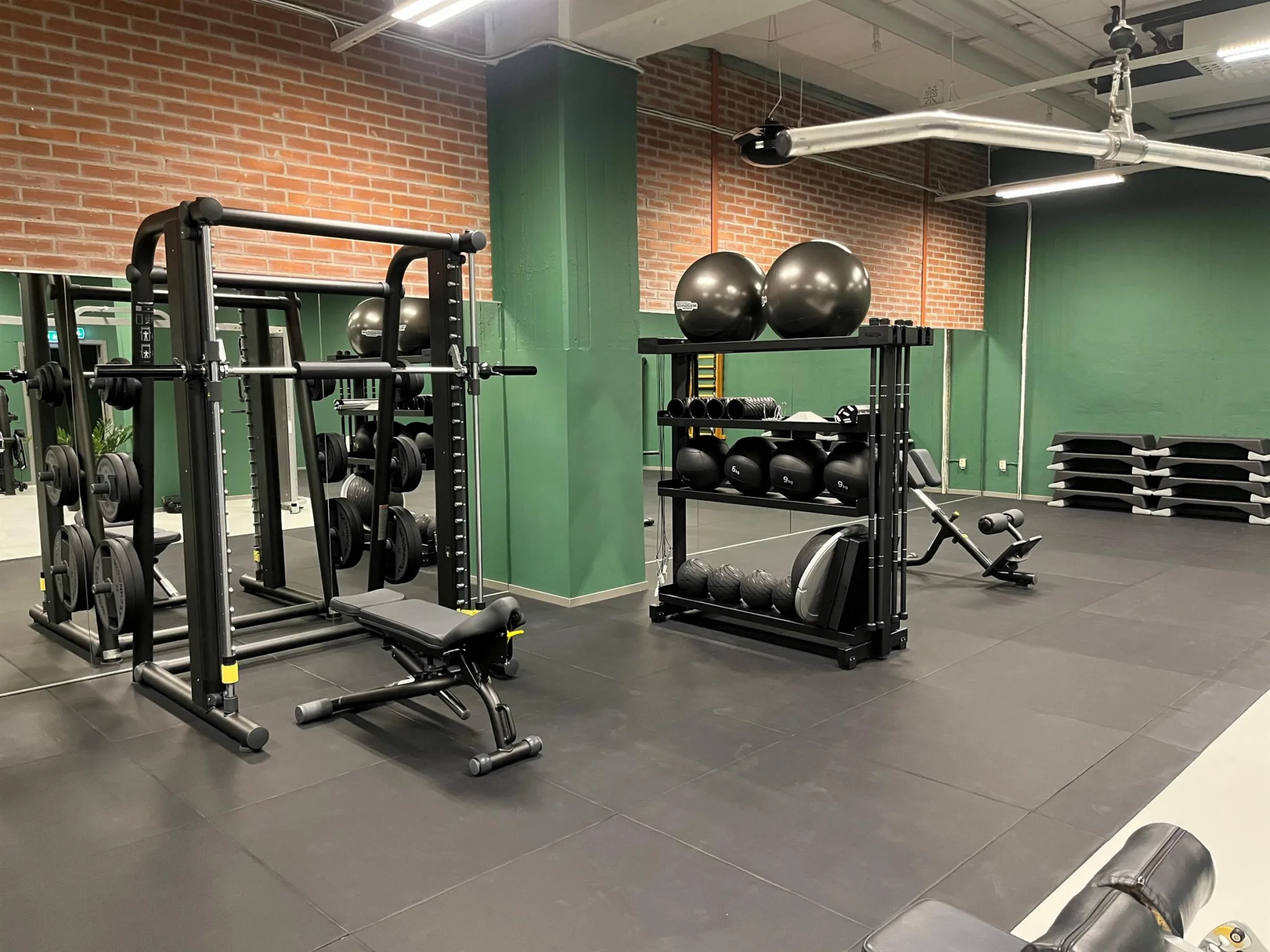 Ett gym med träningsutrustning inklusive skivstänger, vikter och balansbollar, med gröna väggar och tegelväggar.