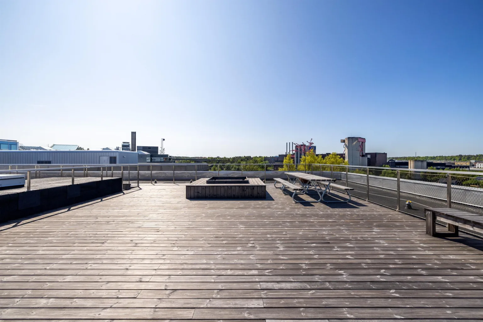 En takterrass med träplankor, bänkar och utsikt över stadens byggnader under en klarblå himmel.