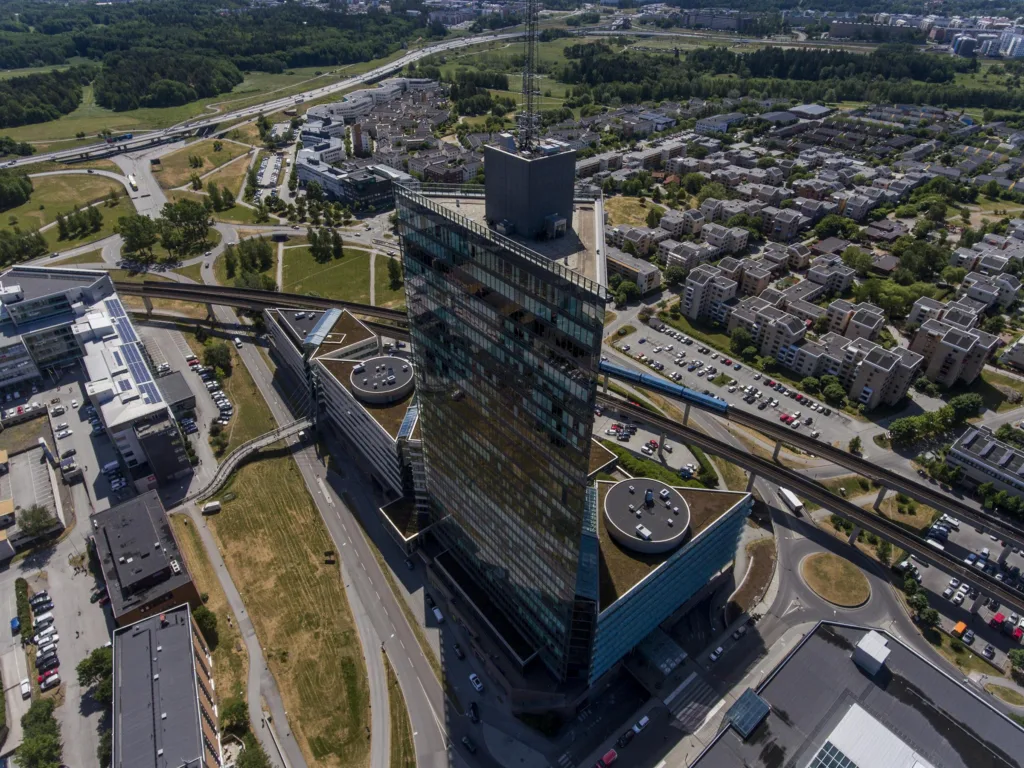 En hög byggnad med glasfasad omgiven av vägar, grönområden och stadslandskap, fotograferad från ovan.