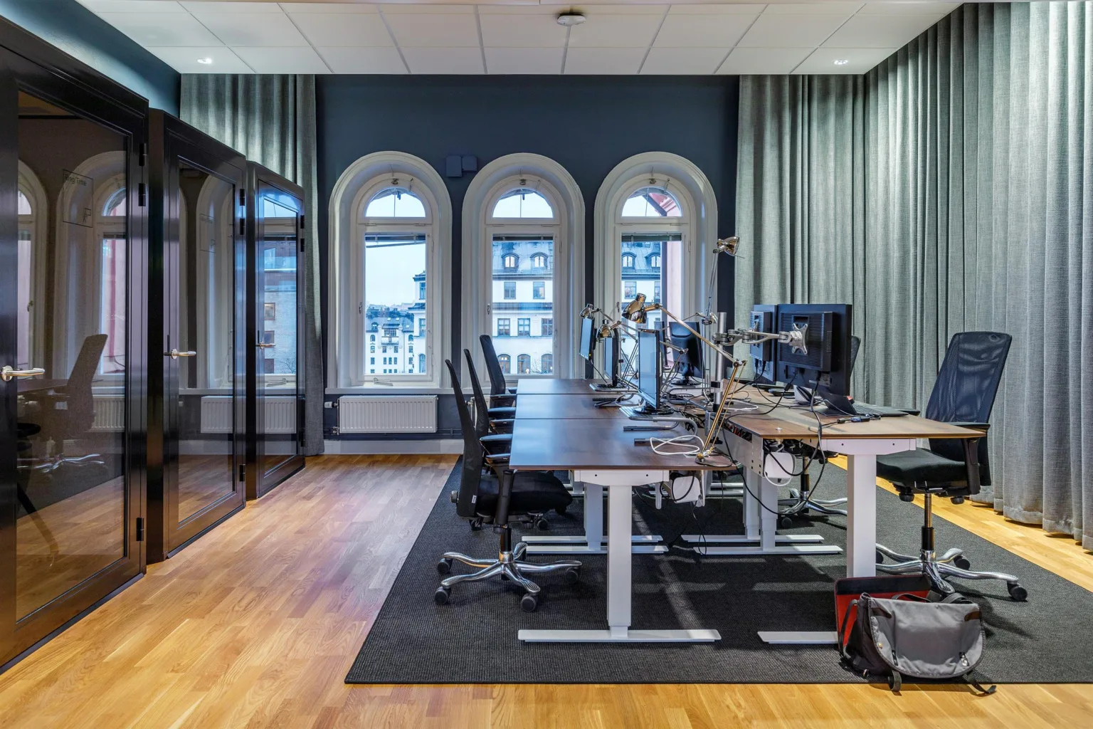 Ett kontorsrum med flera skrivbord och datorskärmar, stora välvda fönster och träparkettgolv.