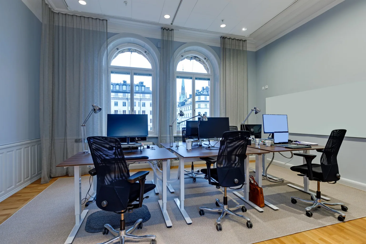 Ett kontorsrum med skrivbord, datorer och skrivbordsstolar framför stora fönster med utsikt över staden.