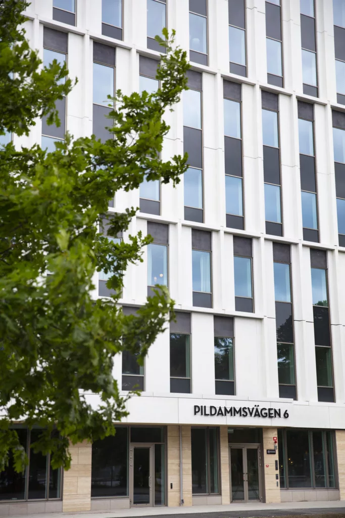 Byggnad med stora glasfönster och texten "PILDAMMSVÄGEN 6" på fasaden, med grönskande träd i förgrunden.