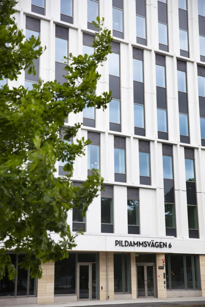 - En modern vit byggnad med stora fönster och texten "Pildammsvägen 6" på fasaden, omgiven av grönska.