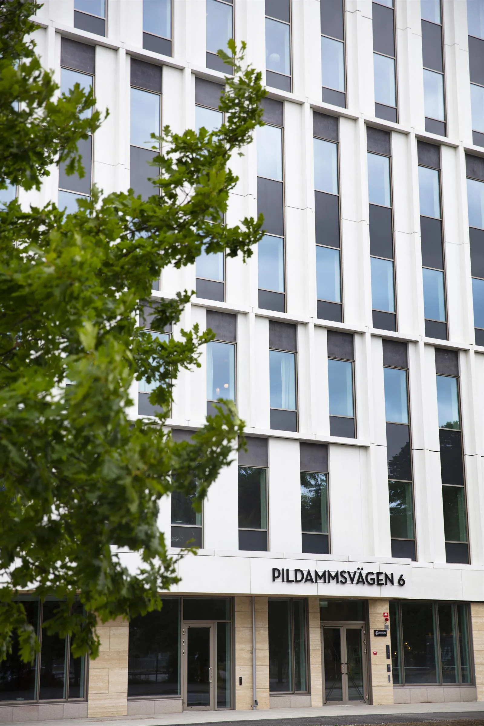 - En modern vit byggnad med stora fönster och texten "Pildammsvägen 6" på fasaden, omgiven av grönska.