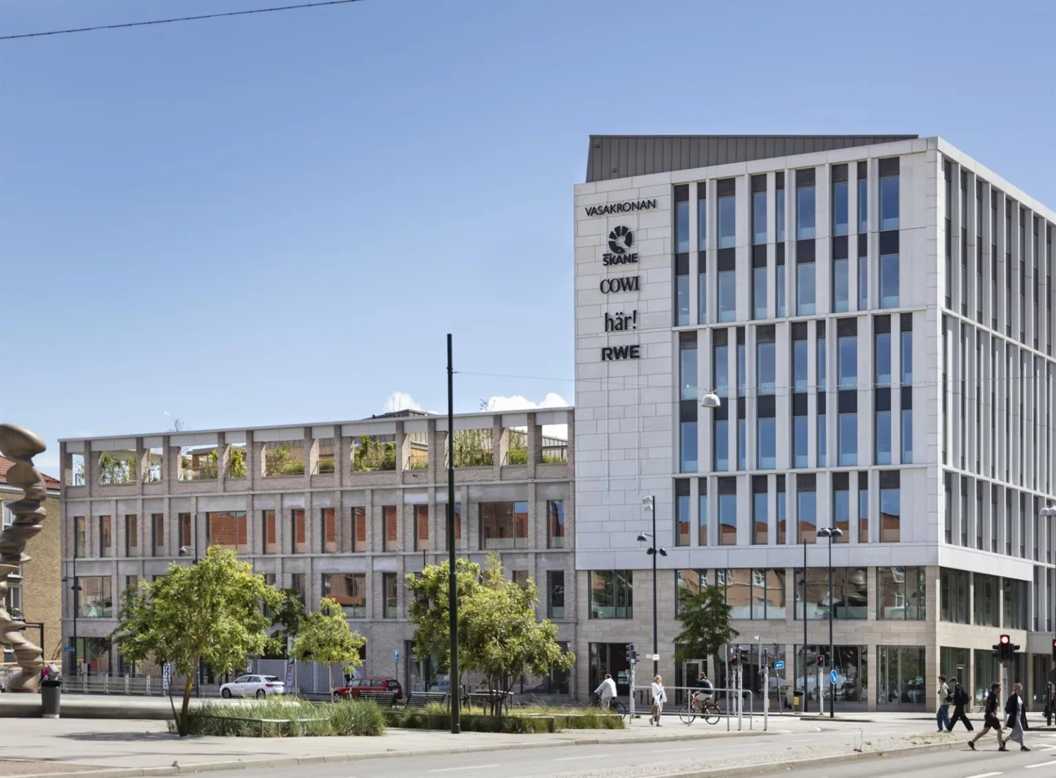 En stor byggnad med flera våningar och glasfönster, omgiven av träd och en blå himmel.