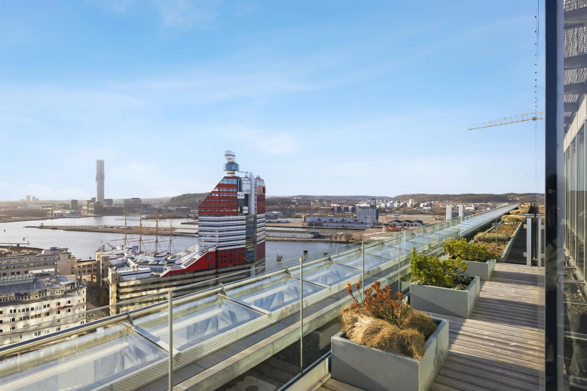 En takterrass med glasräcke, växter och utsikt över en rödvit byggnad.
