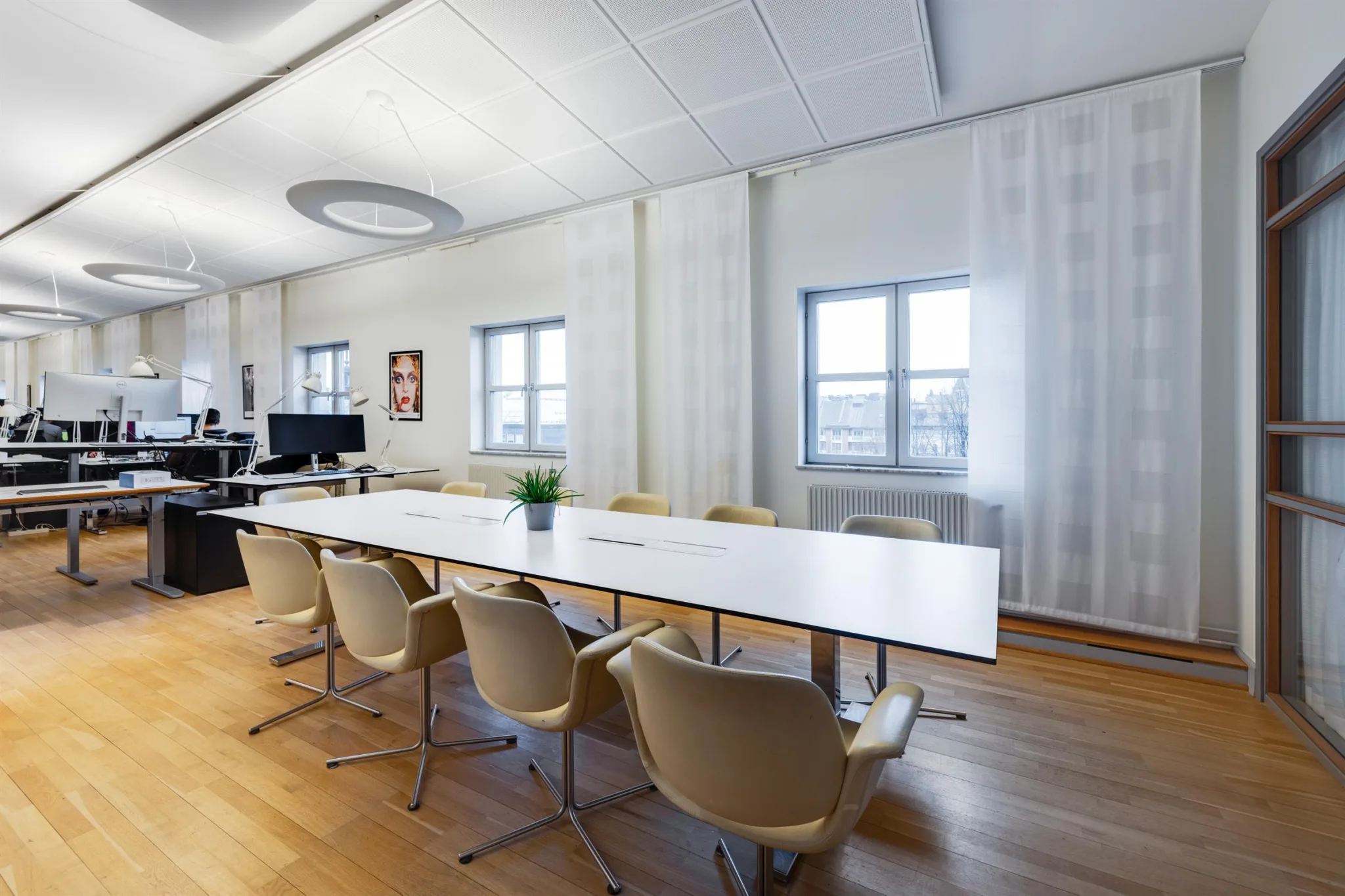 Ett modernt konferensrum med ett långt vitt bord, beige stolar och två fönster som släpper in naturligt ljus.