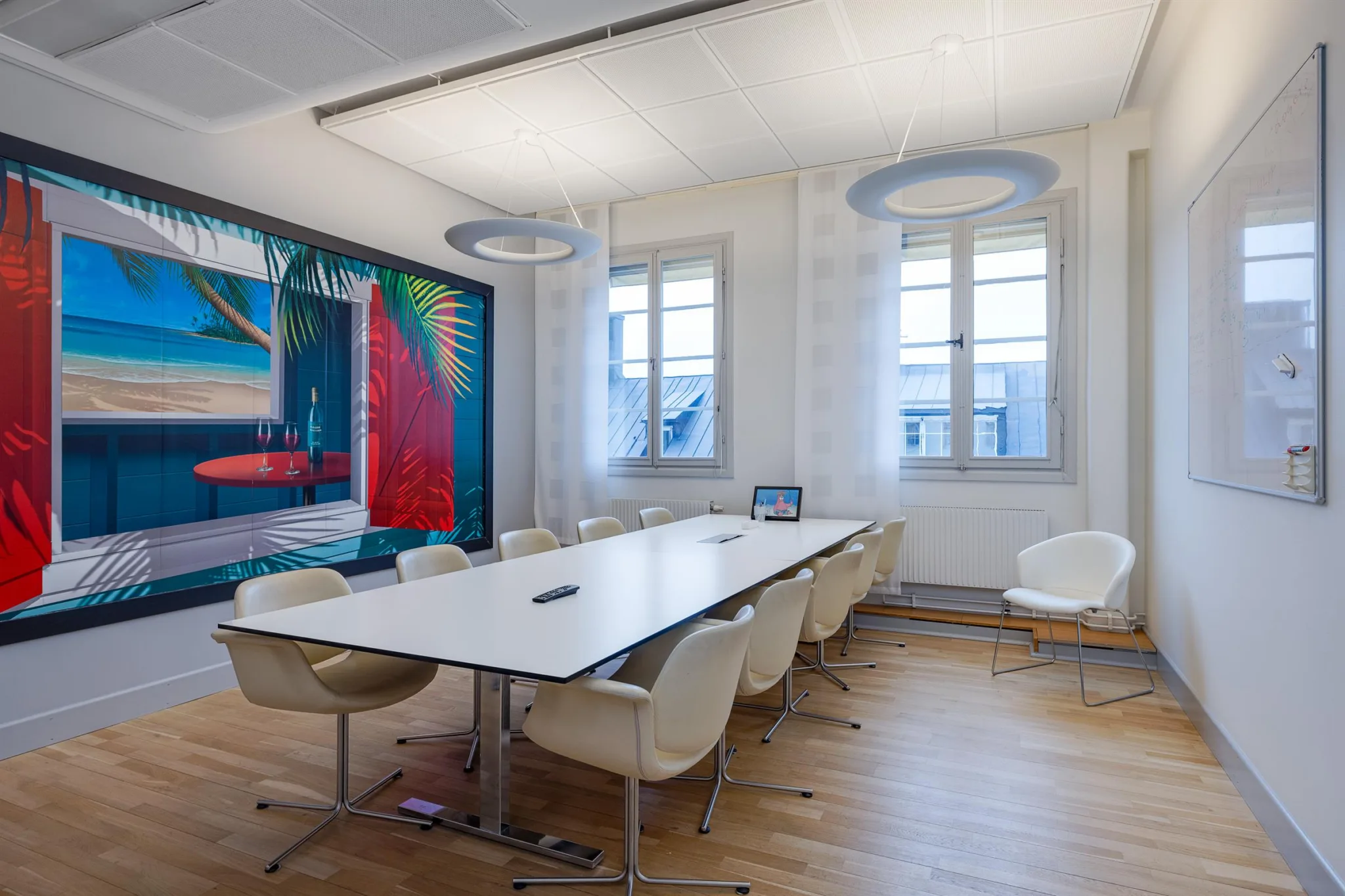 Ett konferensrum med ett stort vitt bord, beige stolar och en färgglad tavla på väggen bredvid två fönster.