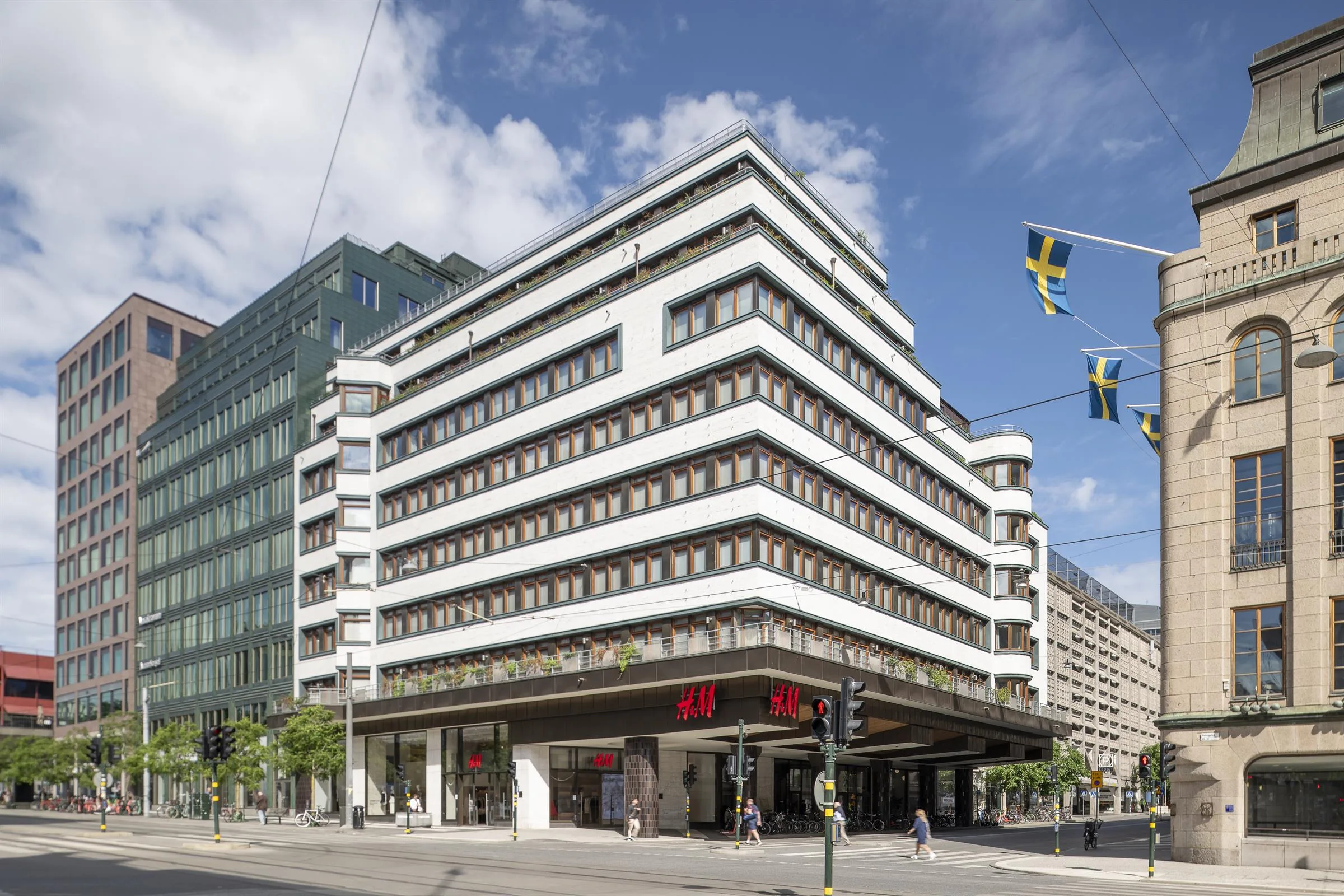 En vit modern byggnad med flera våningar, svenska flaggor hänger över gatan.