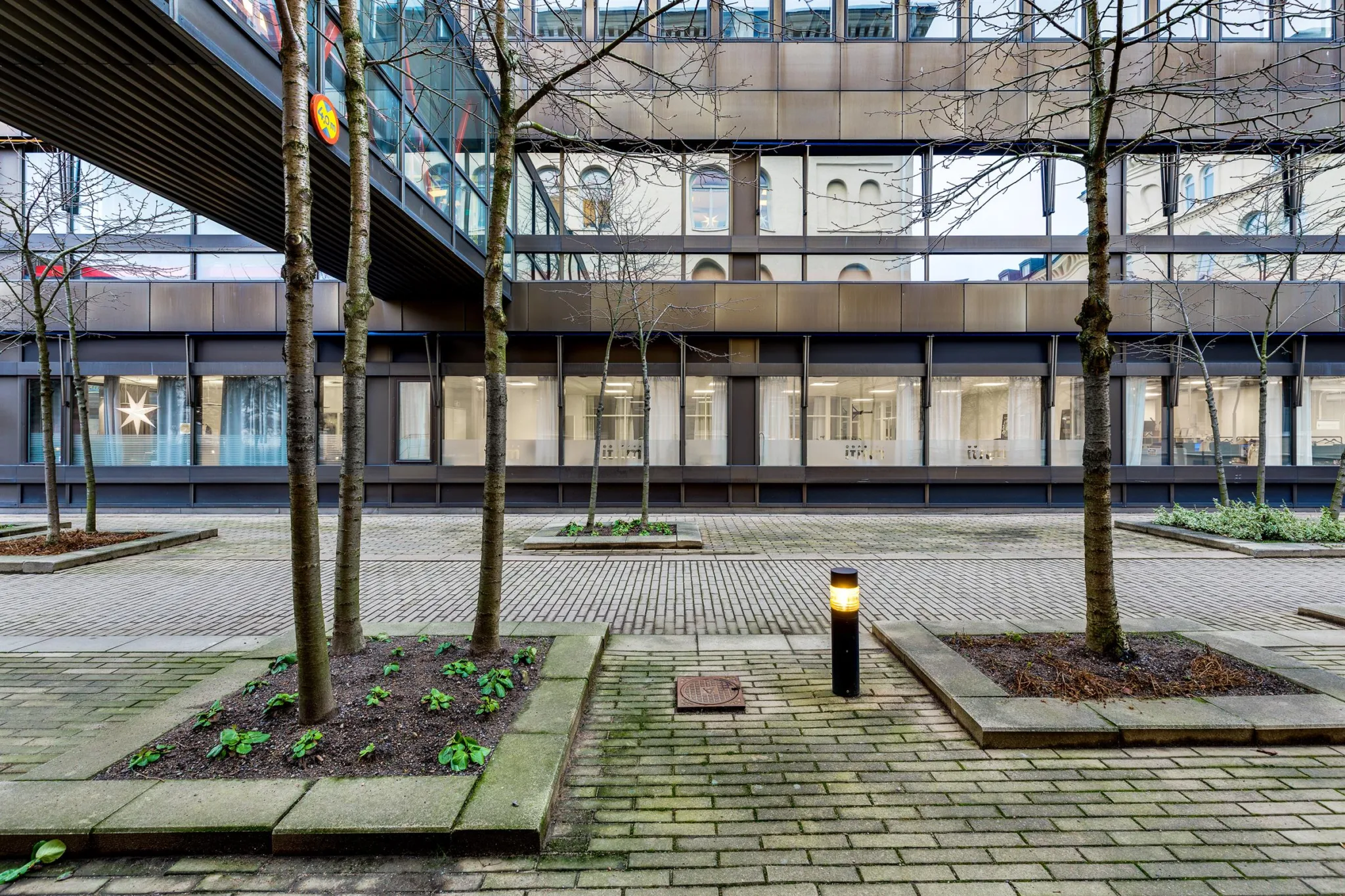 Innergård med träd, belysningsstolpar och stenlagda gångar framför en modern byggnad.