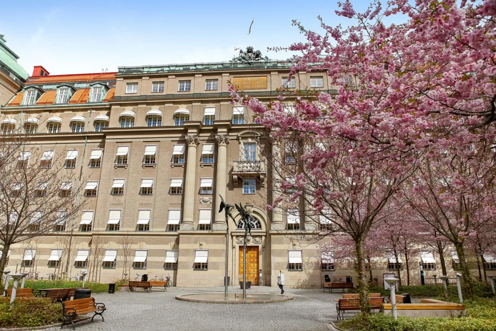 En stor byggnad med körsbärsträd i blom vid en innergård och en gyllene dörr i mitten.