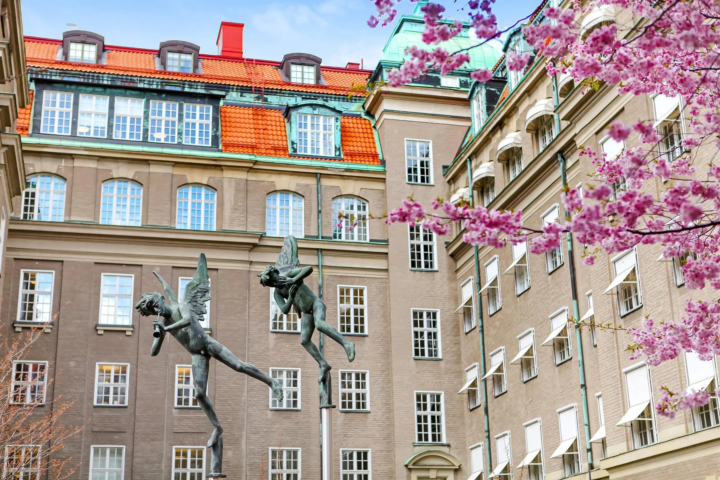 En staty med två dansande figurer framför en byggnad med färgglada tak, omgiven av rosa blommande träd.