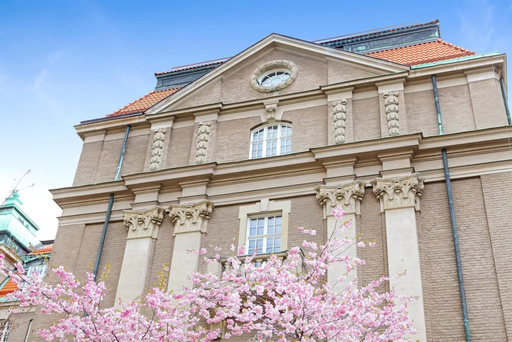 En byggnad med klassisk arkitektur och rosa blommande träd framför, under en klar blå himmel.