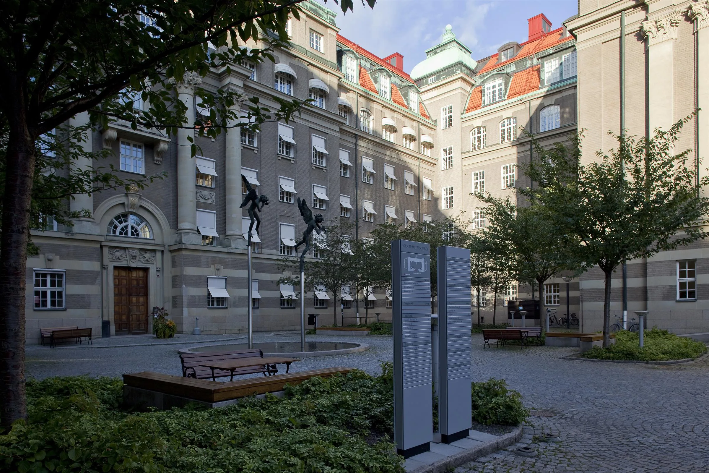 En innergård med en stor byggnad i bakgrunden, en staty och informationsskyltar i förgrunden.