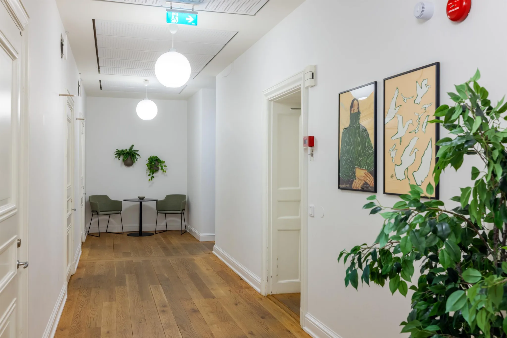 Ljus korridor med trägolv, väggkonst, taklampor och en sittplats med bord och stolar längst bort.