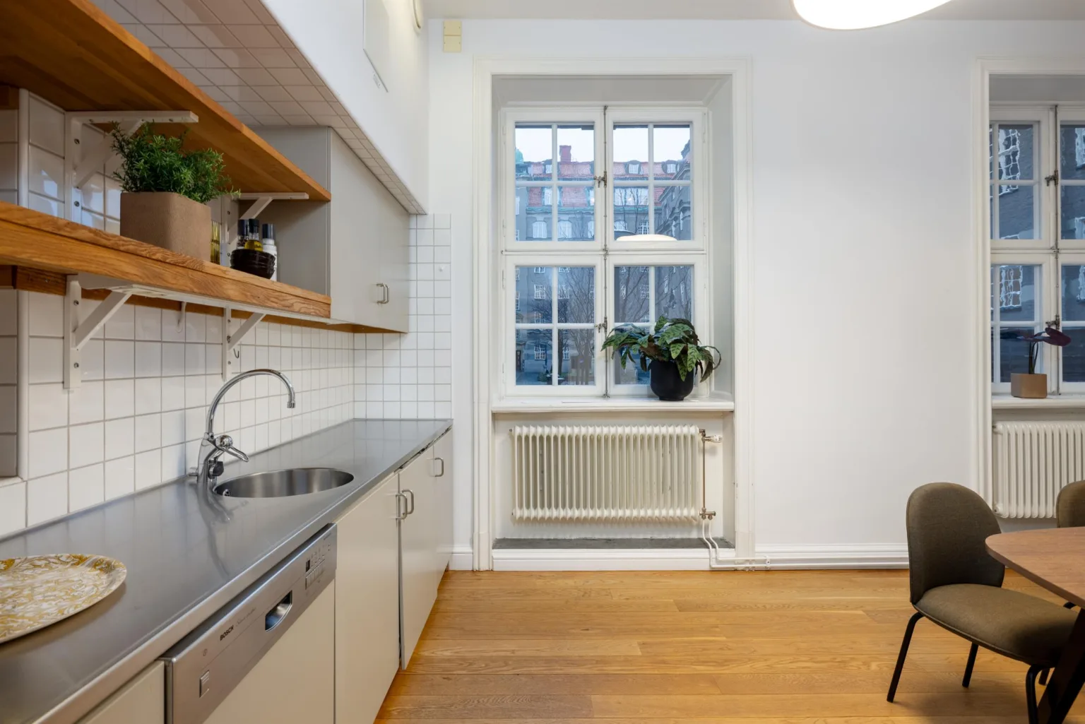 Ljust kök med diskbänk, skåp, hyllor med växter, och ett fönster som släpper in dagsljus.