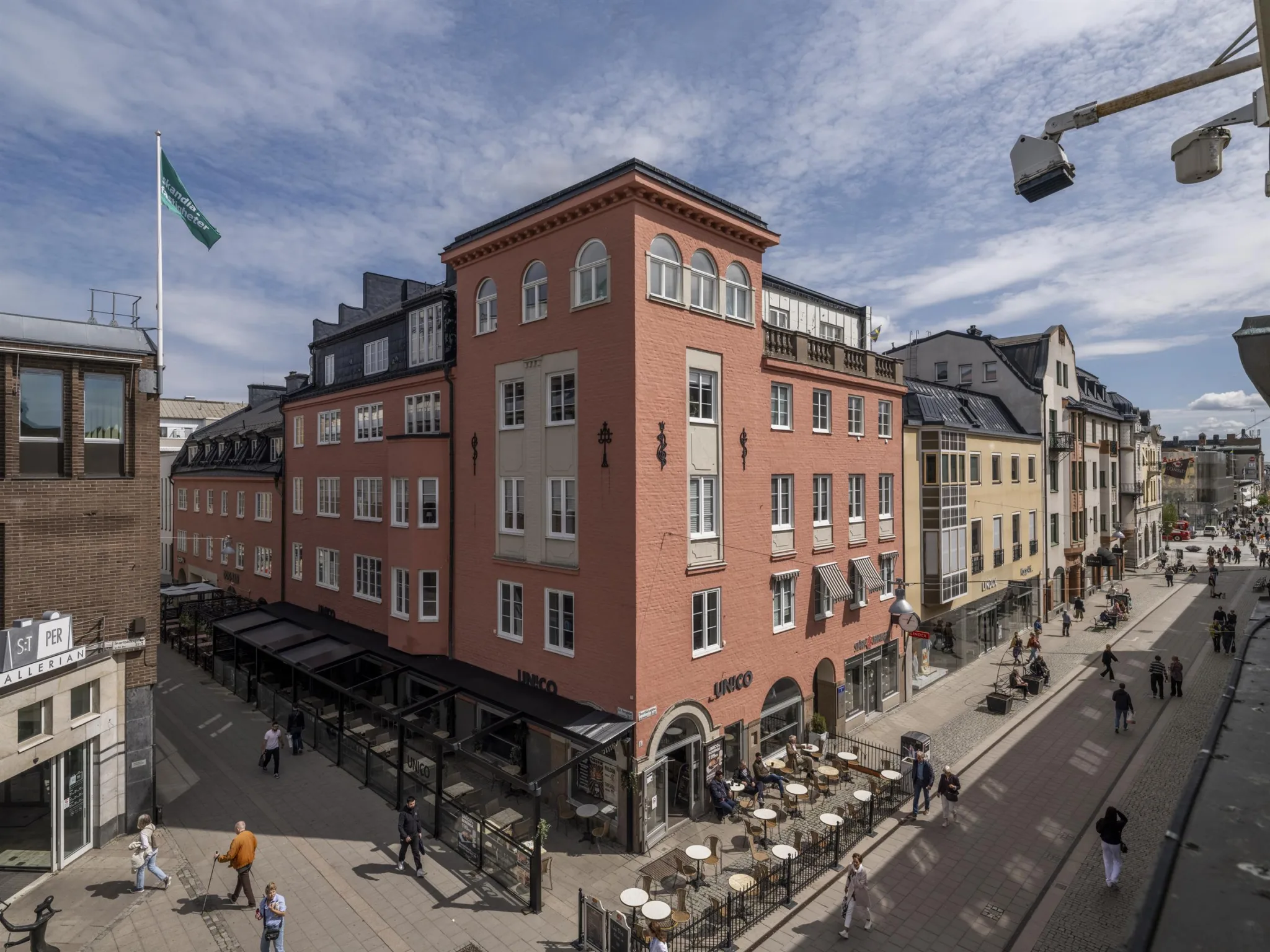 En stadsmiljö med en röd tegelbyggnad med stora fönster och uteserveringar, omgiven av gående människor och blå himmel.