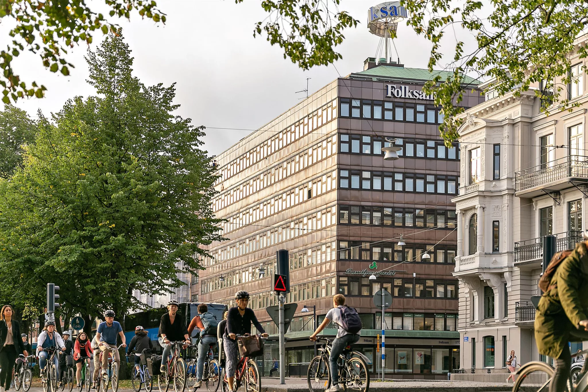 Folk passerar på cyklar framför en stor byggnad där det står "Folksam" högst upp, med träd och trafikljus i förgrunden.
