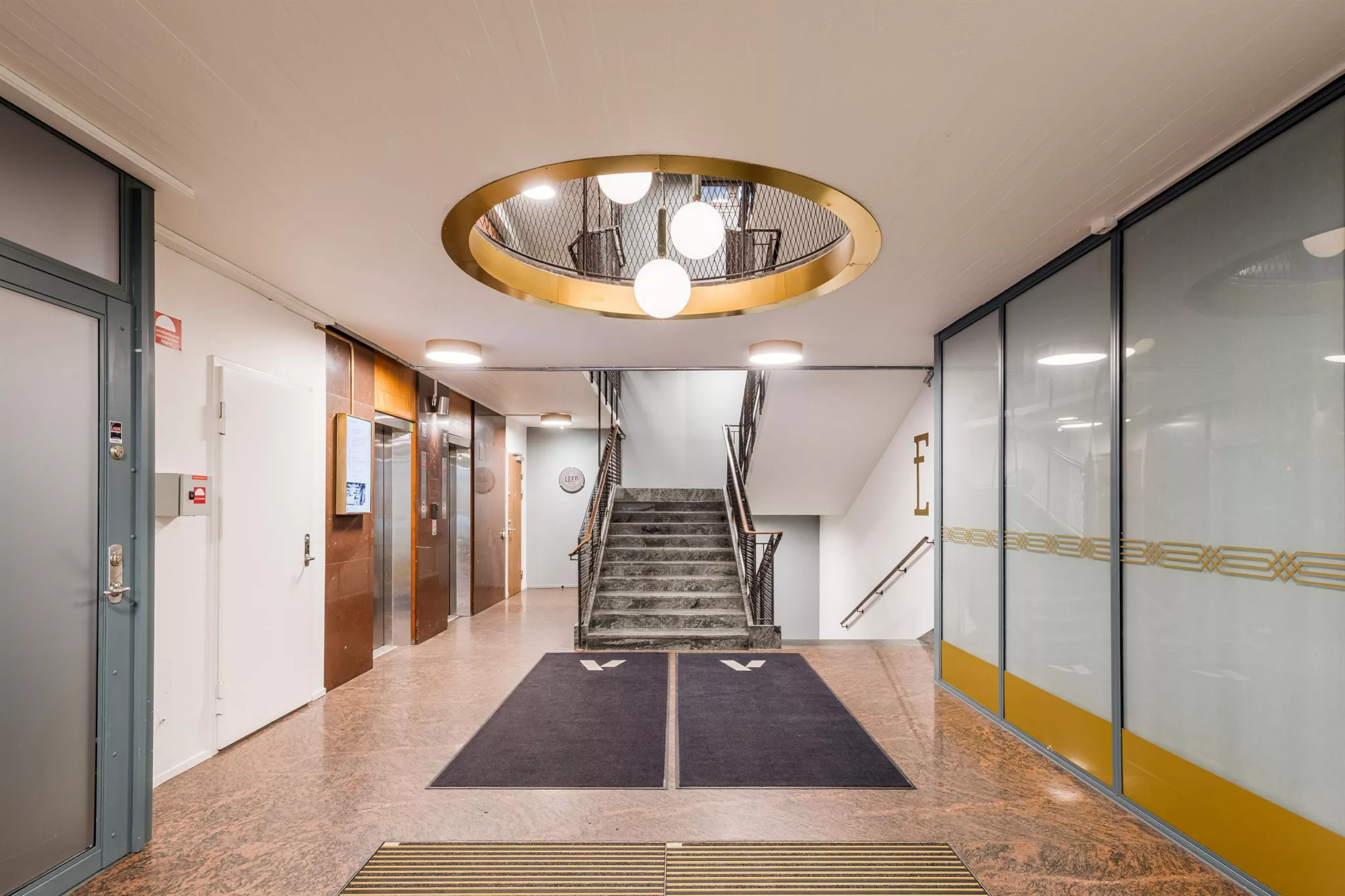 Interiör av en modern entré med trappor, glasväggar och cirkulär takbelysning med flera lampor.