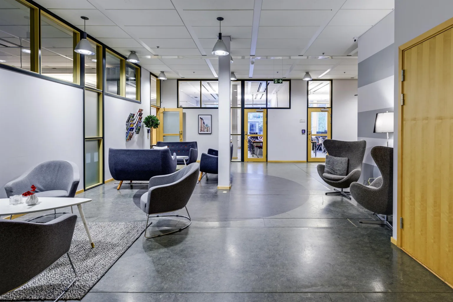 En modern lounge med fåtöljer, soffor och glasväggar som delar utrymmet.