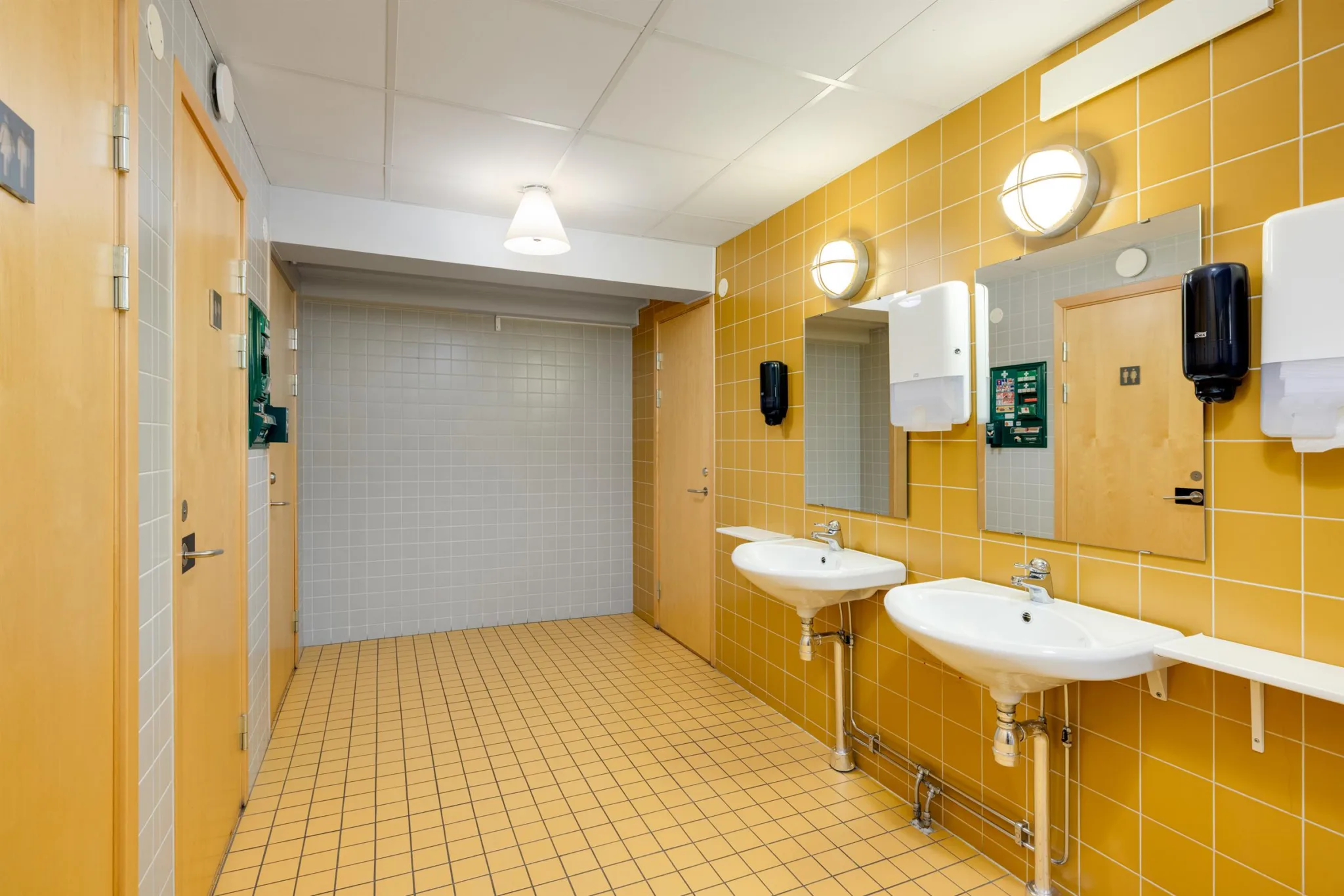 1. Två handfat med speglar och ljusgul kaklad vägg i ett offentligt badrum.