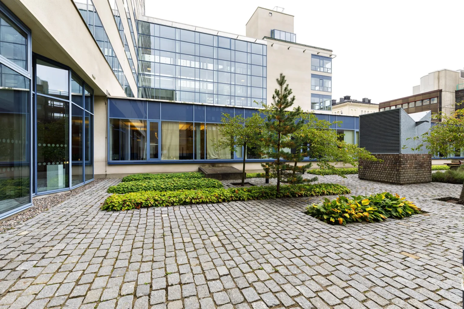 5. En stenlagd innergård med grönska, träd och en modern byggnad omgiven av glasväggar.