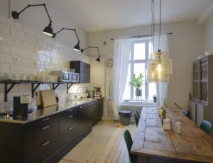 3. Ett kök med mörka skåp, träbord, hängande lampor och ett stort fönster som släpper in dagsljus.