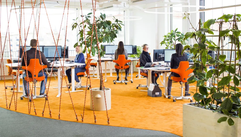 2. Ett kontor med orange matta, gröna växter och personer som arbetar vid skrivbord med datorer.