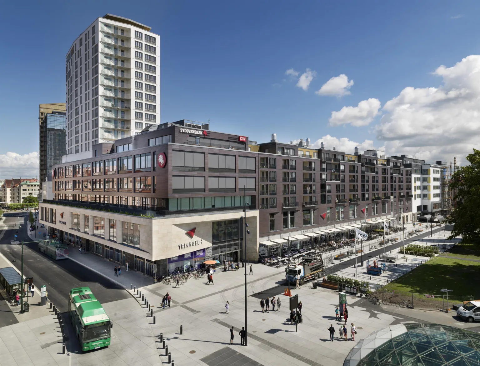 2. Ett stadstorg med höga byggnader, butiker och spårvagn i rörelse under en blå himmel.