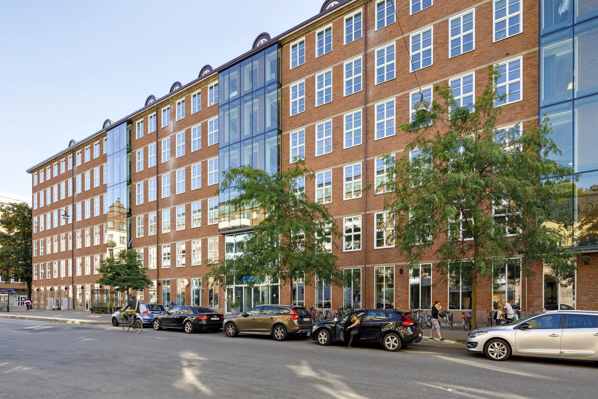 En tegelbyggnad med stora fönster, parkerade bilar och träd längs trottoaren.