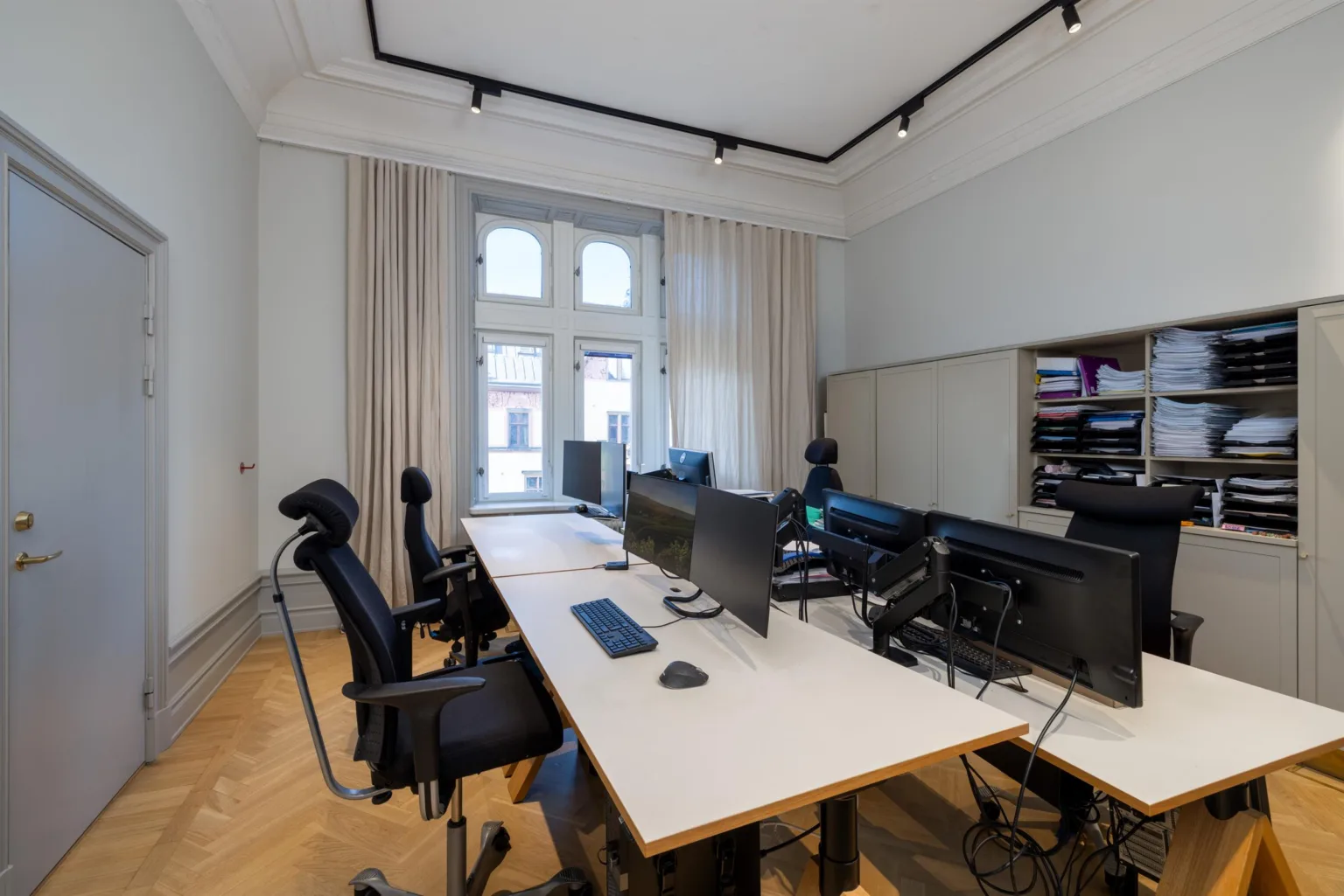 Ett kontorsrum med flera arbetsbord, datorskärmar, ergonomiska stolar och stora fönster med ljusa gardiner.
