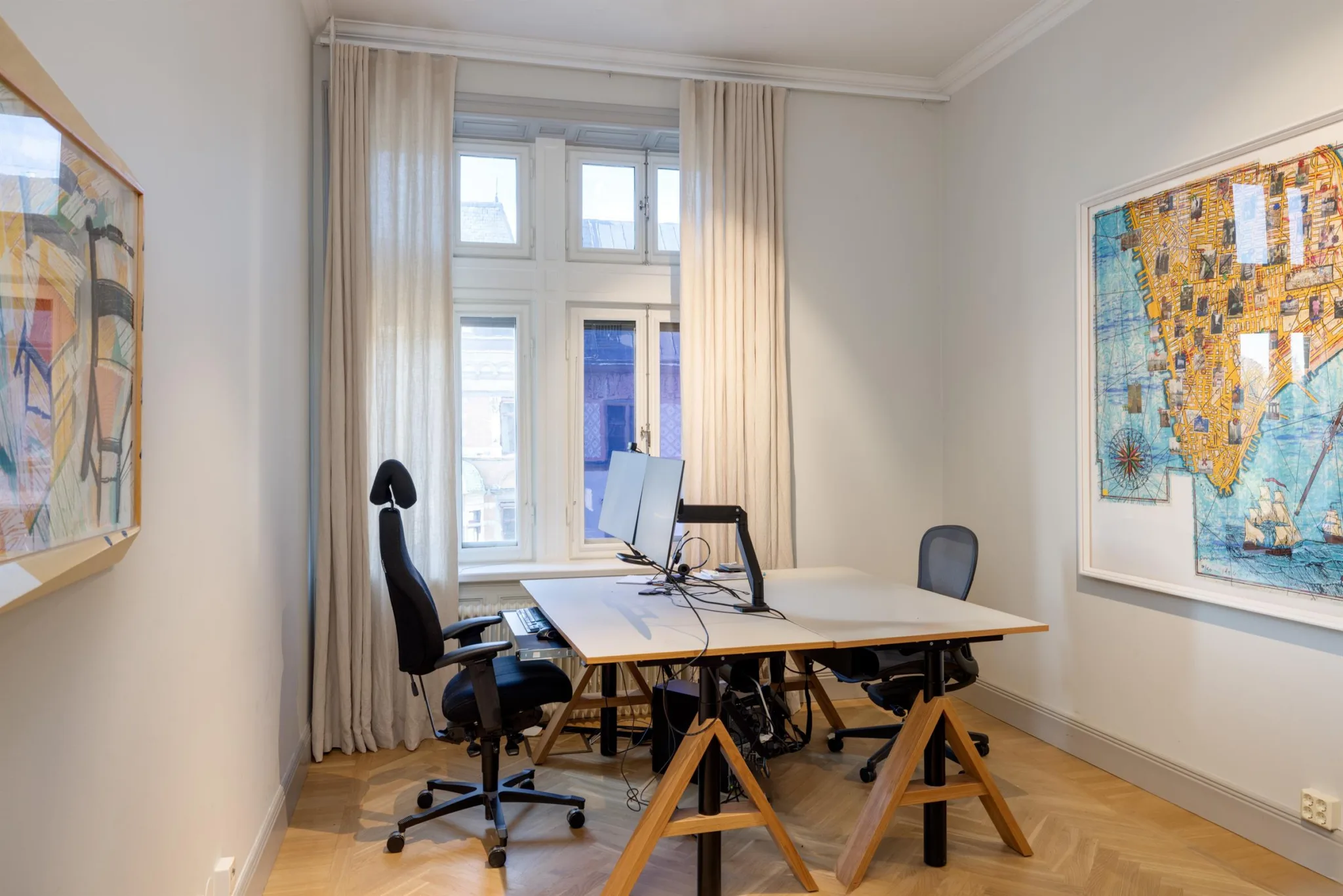 Ett ljust kontorsrum med två skrivbord, ergonomiska stolar och datorer, framför ett stort fönster med vita gardiner.