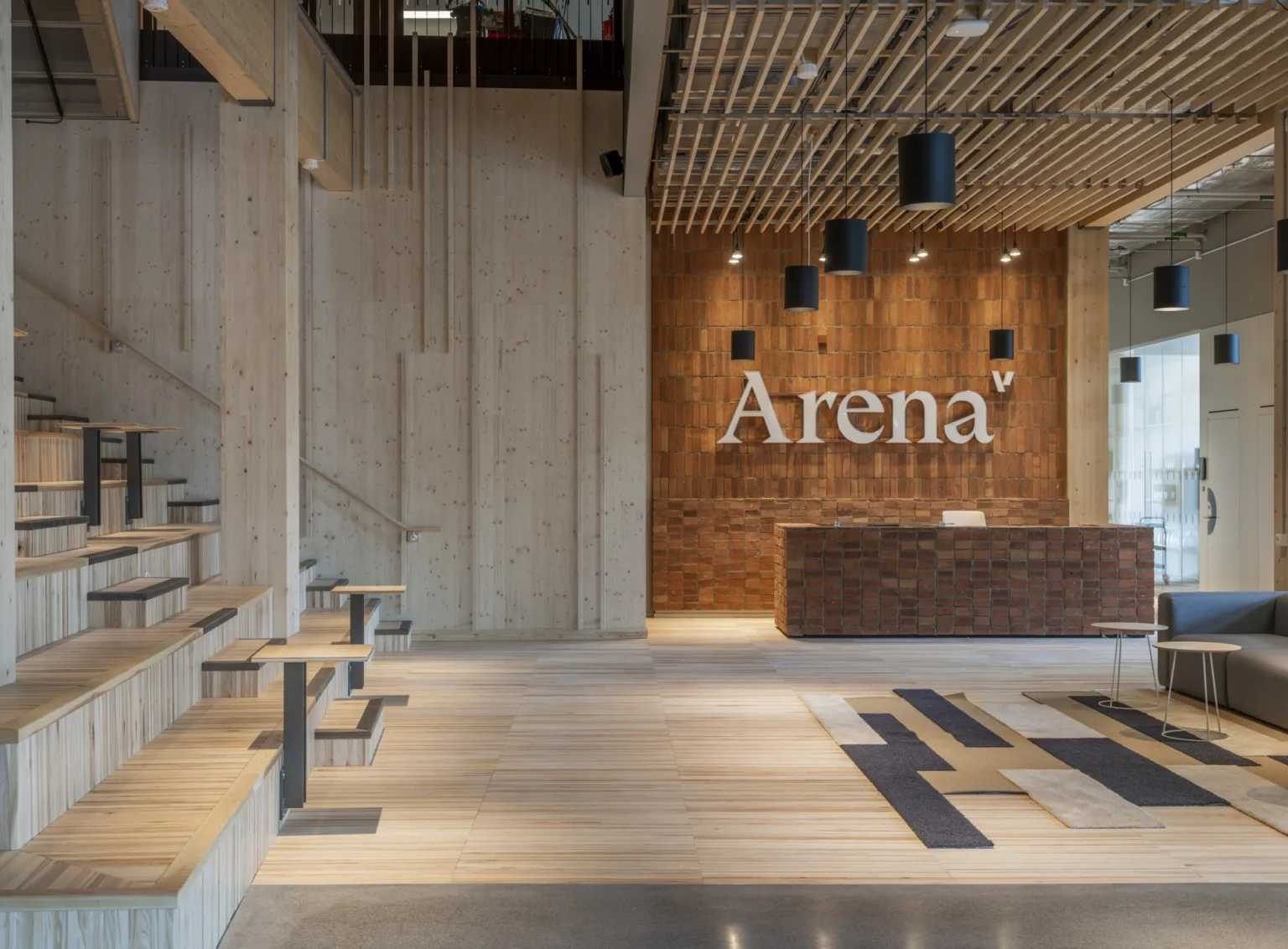 En receptionsyta med trädetaljer och texten "Arena" på en tegelvägg, omgiven av modern inredning och hängande lampor.