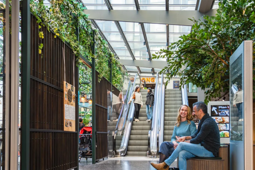 4. Ett inomhuscentrum med gröna växter, rulltrappor och människor som sitter och pratar i en ljus miljö.